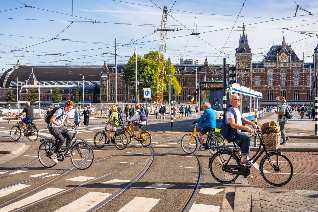 Syklister i sentrum av Amsterdam som krysser et veikryss foran en trikk, med byens hovedjernbanestasjon i bakgrunnen.